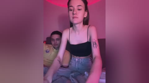 Media: Video of a young, pale-skinned woman with long brown hair in a black crop top and high-waisted jeans, sitting on a bed with a man behind her, both under pink lighting.