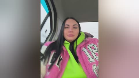 Video of a young woman with long black hair, wearing a bright pink and green jacket with \"18\" on it, inside a car with a blue tinted window.