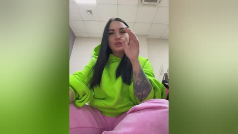 Video of a young woman with long black hair, wearing a neon green hoodie and pink pants, sitting with her hand on her face, in an office with white walls and fluorescent lights.