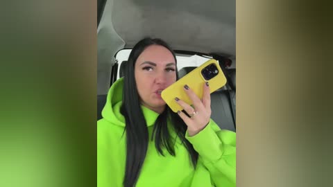 A video of a woman with long black hair, wearing a bright green hoodie, taking a selfie in a car. She holds a yellow phone, lips pursed.
