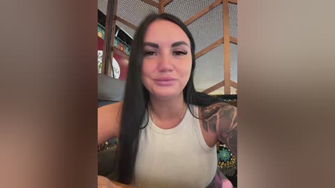 A video of a young woman with long, straight black hair, wearing a white tank top, sitting indoors with a colorful, patterned background.