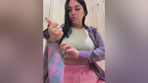 Video of a curvy, light-skinned woman with long black hair, wearing a cream crop top and pink joggers, holding a phone, indoors with a blurred background.