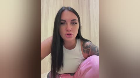 Video of a young woman with long black hair, fair skin, and tattoos, wearing a white tank top, sitting on a pink cushion against a beige curtain backdrop.