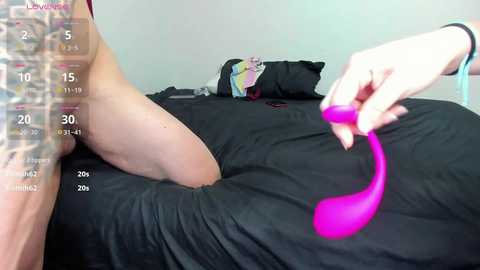 A video of a woman with pale skin, wearing a black bra, lying on a bed. She holds a pink vibrator near her thigh. The room is simple with white walls and black bedding.
