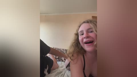 Media: A video of a smiling, fair-skinned woman with curly hair, wearing a black bra, sitting on a bed with patterned sheets. A person, partially out of frame, is brushing her hair.