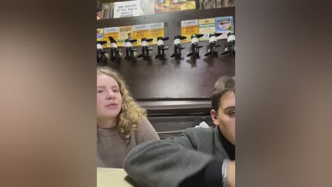Media: Video of two young adults, a blonde woman in a knit sweater and a man with short hair, sitting at a table in a diner. Background shows a counter with black and white statues, and signs.