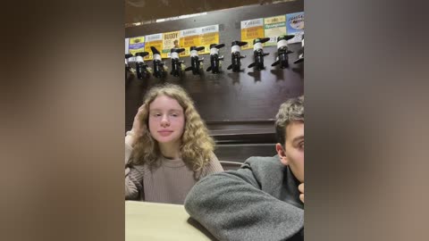 Media: Video of a blonde girl with curly hair, wearing a grey sweater, sitting at a table in a cafeteria, with soda machines in the background.