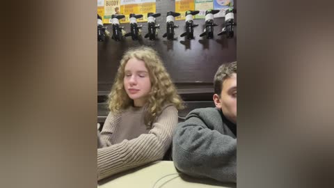 Media: Video of a young blonde girl with curly hair and a boy with short hair, both wearing casual clothes, sitting close in a dimly lit room with a row of black and white beer taps in the background.