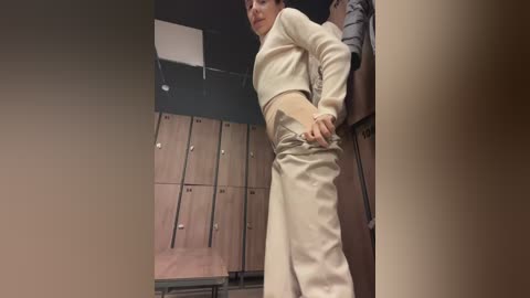 Media: Video of a woman in beige workout attire adjusting her pants in a dimly lit locker room with wooden lockers and a ceiling light.