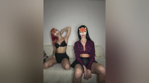 Video of two women with cat masks, one wearing a black bra and panties, the other a plaid shirt, seated on a beige couch.