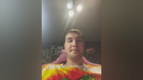 Media: A video of a young, fair-skinned, short-haired boy in a colorful tie-dye shirt, seated in a dimly lit room with a ceiling light, surrounded by scattered books and posters on the wall.