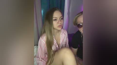 Video of a young woman with long, straight blonde hair, wearing a pink and white striped pajama top, sitting on a bed. A woman with short blonde hair and sunglasses is partially visible behind her. The background features a teal curtain and soft lighting.