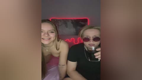 Video of two young women, one topless with pink bra, the other wearing sunglasses, sitting on a red couch in a dimly lit room.
