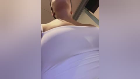 Media: Video of a woman with a round, pale face and short dark hair, wearing a white tank top. The image is taken from a low angle, emphasizing her breasts.