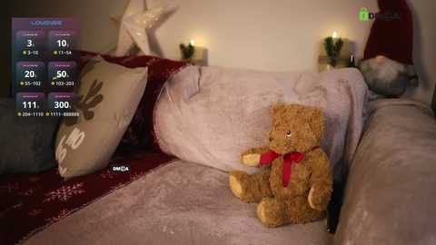 Media: A cozy living room with a plush, cream-colored couch featuring a teddy bear, red scarf, and festive decorations. The background includes two lit candles, a wrapped present, and a snowman ornament.