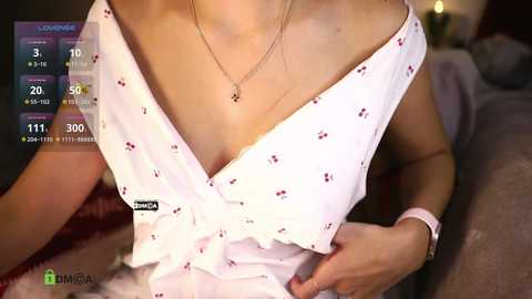 Media: Video of a woman in a white, floral-patterned, off-the-shoulder dress, showcasing a low neckline. She wears a delicate necklace and a wristwatch. Background features a dimly lit room.