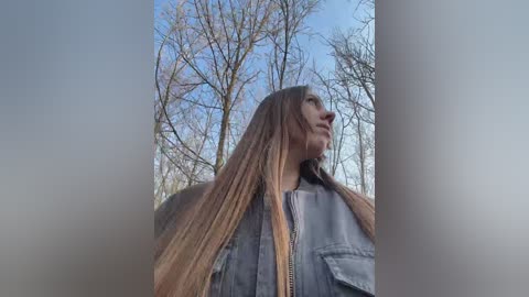 Media: Video of a young woman with long, straight, light brown hair, wearing a denim jacket, standing in a forest with bare trees and a blue sky.