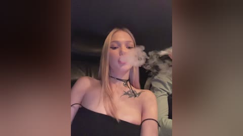 Media: Video of a blonde woman with a black choker and off-shoulder black dress, blowing smoke, in a dimly lit room.