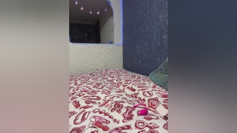 Media: A video of a small, dimly lit bedroom with a white bedspread featuring red, abstract designs. A pink vibrator is placed on the bed, and the walls are textured with light and dark shades of blue.