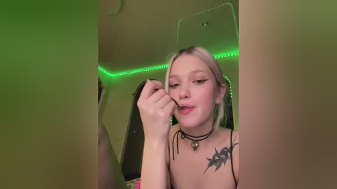 Media: Video of a young woman with blonde hair, fair skin, and tattoos, wearing a black choker and bra, applying lipstick, in a dimly lit room with green LED strip lighting.