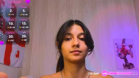 Video of a young woman with dark hair, wearing a pink top, standing against a light wooden wall with a framed artwork in the background. A virtual health tracker overlay shows her stats.