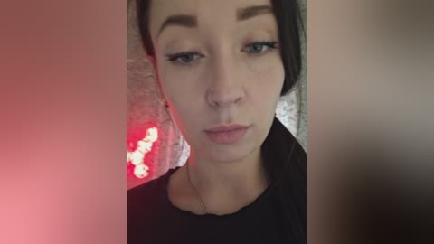 A video of a young woman with fair skin, dark hair, and a serious expression, wearing a black top, against a blurry, textured background with red and white lights.
