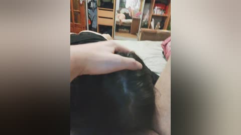 Video of a person lying on a bed, with a hand gently touching their head, in a cluttered bedroom with a wooden wardrobe and shelves visible in the background.