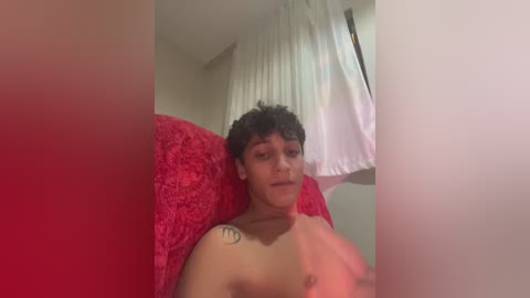 A video of a topless young person with curly hair and a tattoo on their left shoulder, lying on a red fuzzy blanket in a dimly lit room with white curtains.