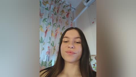 Video of a young Caucasian woman with straight brown hair, smiling slightly, standing in a room with floral-patterned curtains and a white wall.