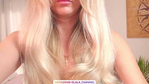 Video of a light-skinned woman with long, platinum blonde hair, wearing a cross necklace, in a cozy room with beige walls and a framed abstract painting.