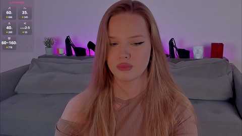 Video of a Caucasian woman with long, straight blonde hair, wearing a beige off-shoulder top, sitting on a grey couch. Background features a purple-lit wall with black high-heeled shoes, potted plants, and a white lamp.