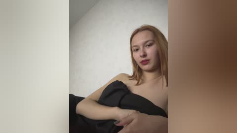 Media: Video of a fair-skinned, light-haired woman with red lipstick, holding a black cloth against her chest, against a plain white wall, in a dimly lit room.