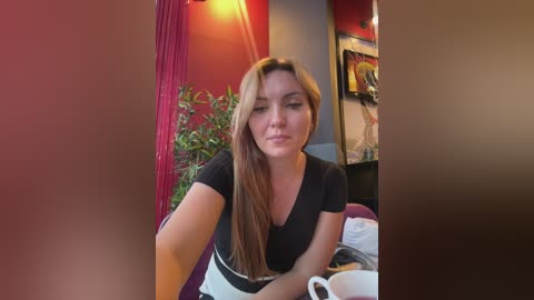 Video of a Caucasian woman with long, straight, light brown hair, wearing a black t-shirt and white apron, sitting at a table in a cozy caf\u00e9 with red curtains, green plants, and colorful artwork on the wall.