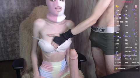Video of a pale, slender woman in a white bra and pastel skirt, with a white mask and black gloves, sitting on a chair, being touched by a man in green Calvin Klein boxers.
