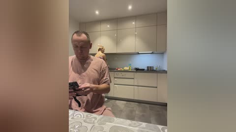 Media: Video of a man in a pink hospital gown, holding a smartphone, in a modern kitchen with white cabinets, a blonde woman in a light-colored top, and a grey countertop.