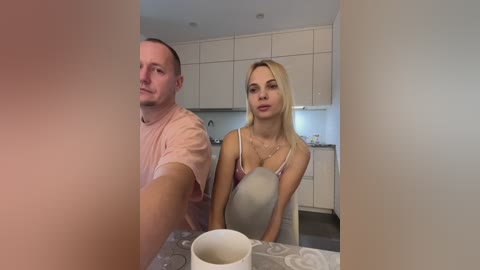 Media: Video of a blonde woman in a pink camisole and a man in a pink shirt, seated at a kitchen table, with a white mug in front.