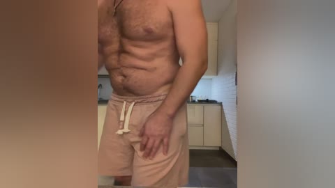 Media: A video of a shirtless, muscular man with light skin, wearing loose beige shorts, standing in a modern kitchen with white cabinets and a backsplash.