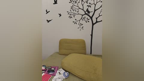 Media: Video of a cozy, minimalist room with beige armchairs, a mustard-yellow cushion, a book and sunglasses on a table, and a black tree wall decal with birds.