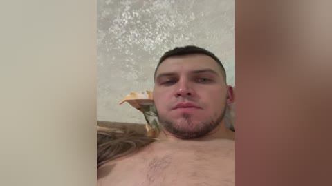 A video of a young man with short dark hair, light skin, and a trimmed beard, lying shirtless against a textured, off-white wall, partially obstructed by a vertical divider.