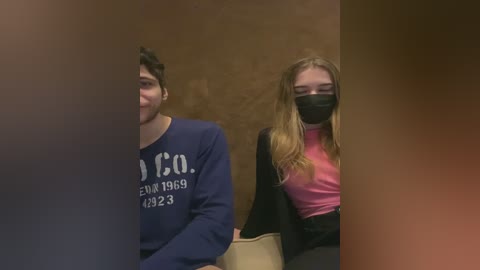 Video of two people in a dimly lit room, a man with short dark hair in a navy hoodie, and a woman with long blonde hair in a black mask and pink top.