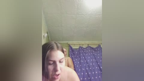 Video of a woman with light skin and straight, shoulder-length brown hair, partially seen from the side. She's topless, standing in a small, dimly lit bathroom with a textured ceiling and a purple shower curtain adorned with white polka dots.