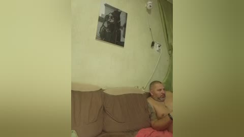 Video of a bald, muscular man with tattoos, shirtless, lying on a bed covered in beige sheets. A black-and-white framed photo hangs on a yellow wall.