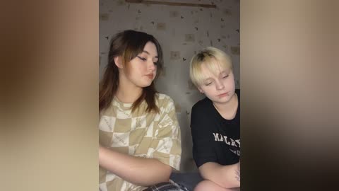 Media: Video of two young women, one with long dark hair and patterned shirt, the other with short blonde hair and black shirt, seated in a dimly lit room with floral wallpaper.