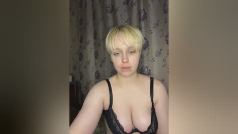 Media: A video of a fair-skinned woman with short blonde hair, wearing a black lace bra, standing in a dimly lit room with floral-patterned curtains.