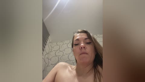 Video of a young, nude, fair-skinned woman with long brown hair, looking down with a neutral expression. The background features a beige wall with a patterned wallpaper.