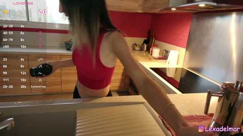 Video of a slim, fair-skinned woman in a red crop top and black leggings, using a controller in a modern kitchen with wooden cabinets and red walls.