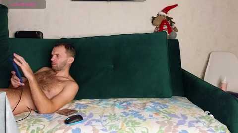 Video of a shirtless, bearded man with short hair, playing a video game on a green couch with a colorful blanket. In the background, a Christmas decoration and a stuffed animal are visible.