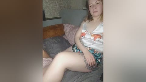 A candid video shows a fair-skinned woman with shoulder-length blonde hair, wearing a white camisole with fox prints and floral shorts, reclining on a bed with gray bedding.