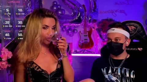 Media: Video of a blonde woman in a black lace dress and a masked man in a black t-shirt, sitting in a dimly-lit room with musical instruments, a guitar, and pink balloons.