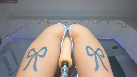 Video of a woman's bare legs and buttocks, featuring a large, tan vibrator strapped between her thighs, with blue ribbon tattoos on her inner thighs.